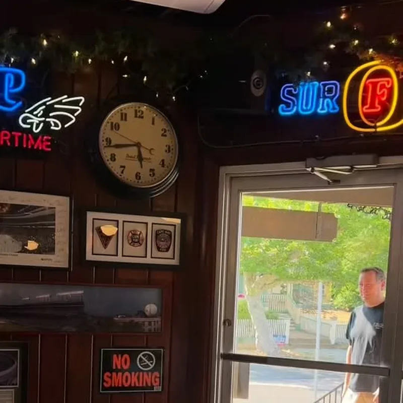 Inside the C-View: dark wood panelled walls, an Eagles Overtime neon and a Surf neon, a schoolhouse clock, framed prints and a No Smoking sign, and the front door standing open onto the sunlit street