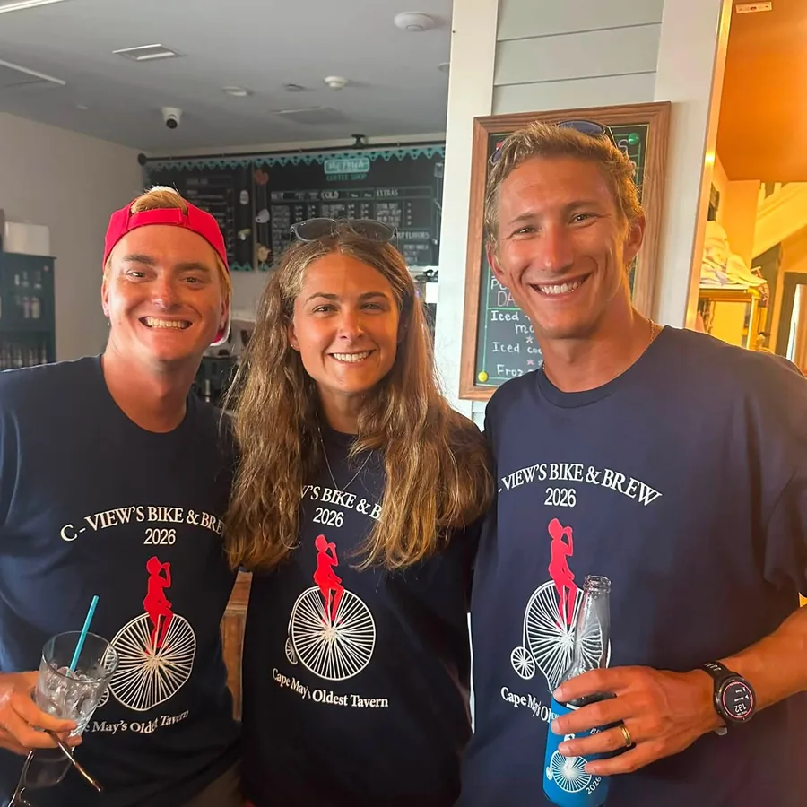 Three riders in navy C-View’s Bike & Brew 2026 shirts, printed with a penny-farthing and the line Cape May’s Oldest Tavern, holding drinks at a bar