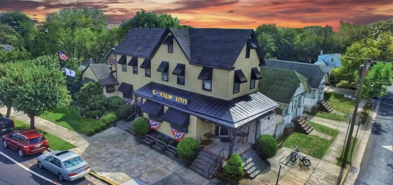 The C-View Inn from the air at sunset: a three-storey yellow clapboard building on the corner, with a black awning lettered C-VIEW INN, red white and blue bunting along the porch, and benches and a bike rack on the sidewalk